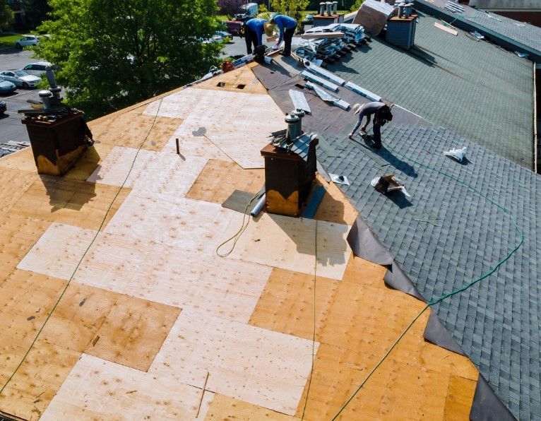 Roofers installing new shingles on a building roof, with wooden panels, chimneys, and parked cars nearby.
