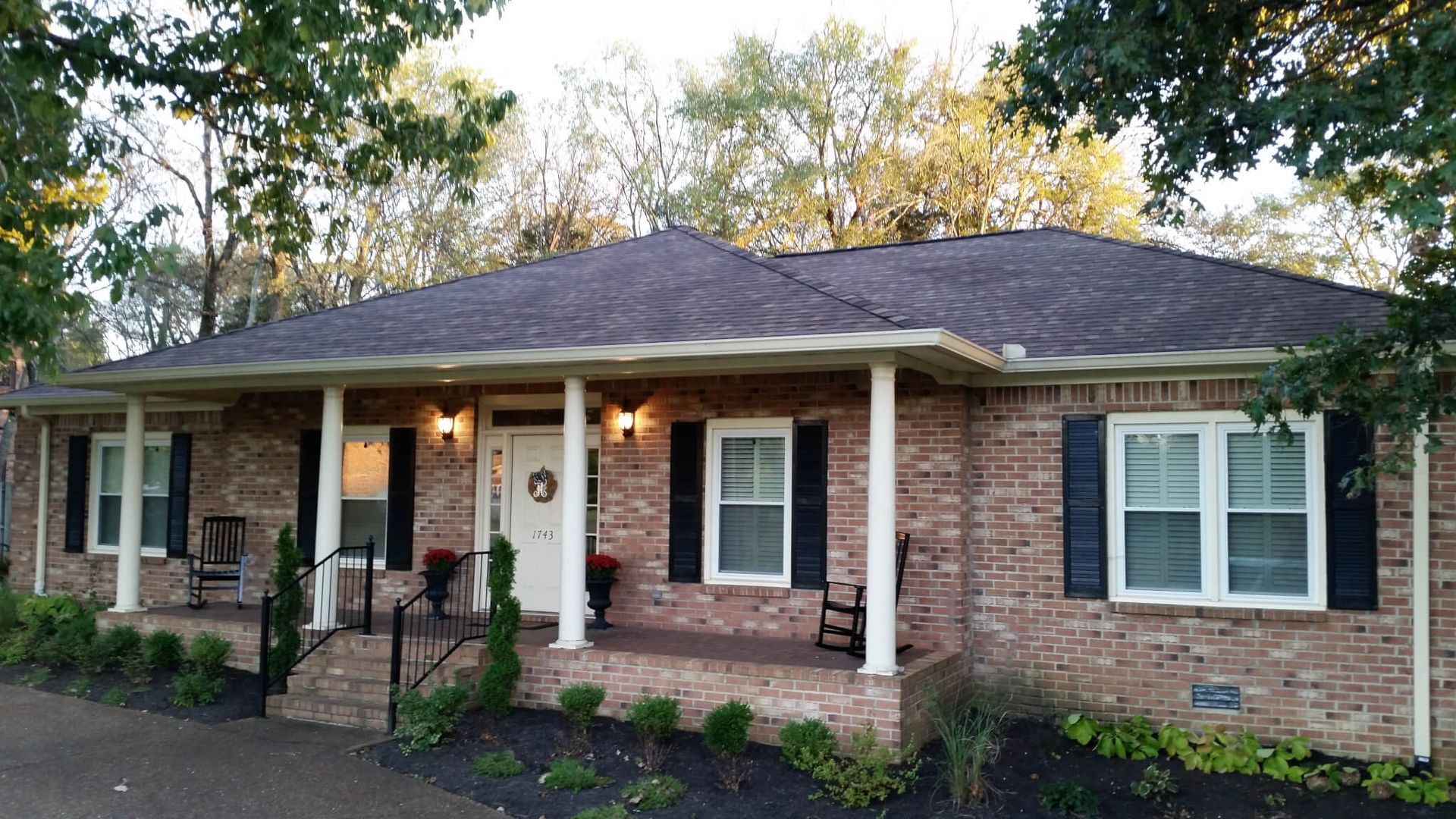 Brick ranch-style house with dark roof and porch, surrounded by trees.
