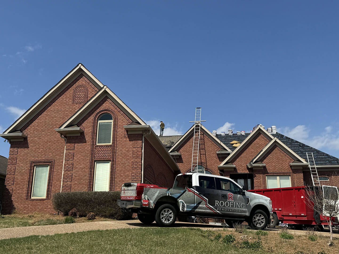 A red brick house with a roofing truck and a dumpster on the lawn under a clear blue sky.