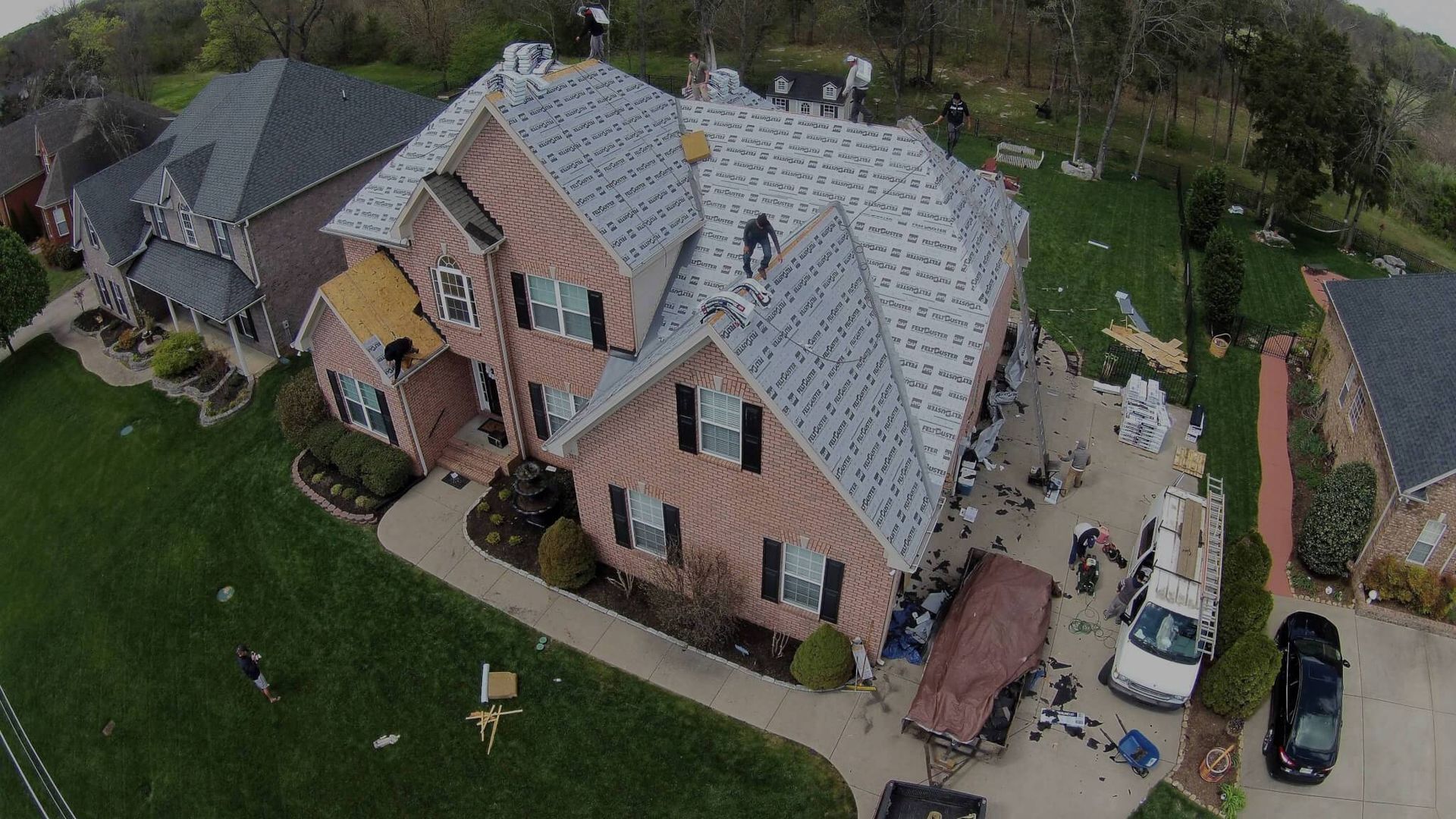 Aerial view of a house with roofing being replaced. Workers, materials, and vehicles are visible in the driveway.