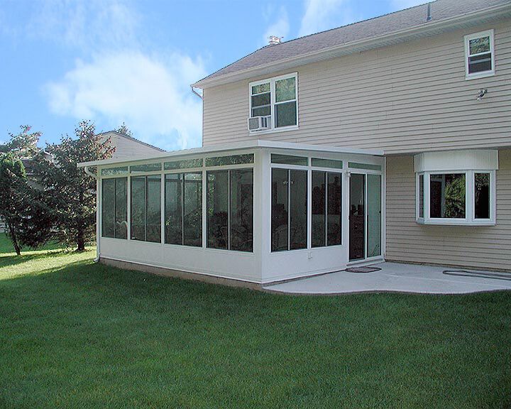 White sunroom addition to a beige house with a concrete patio and green lawn on a sunny day.