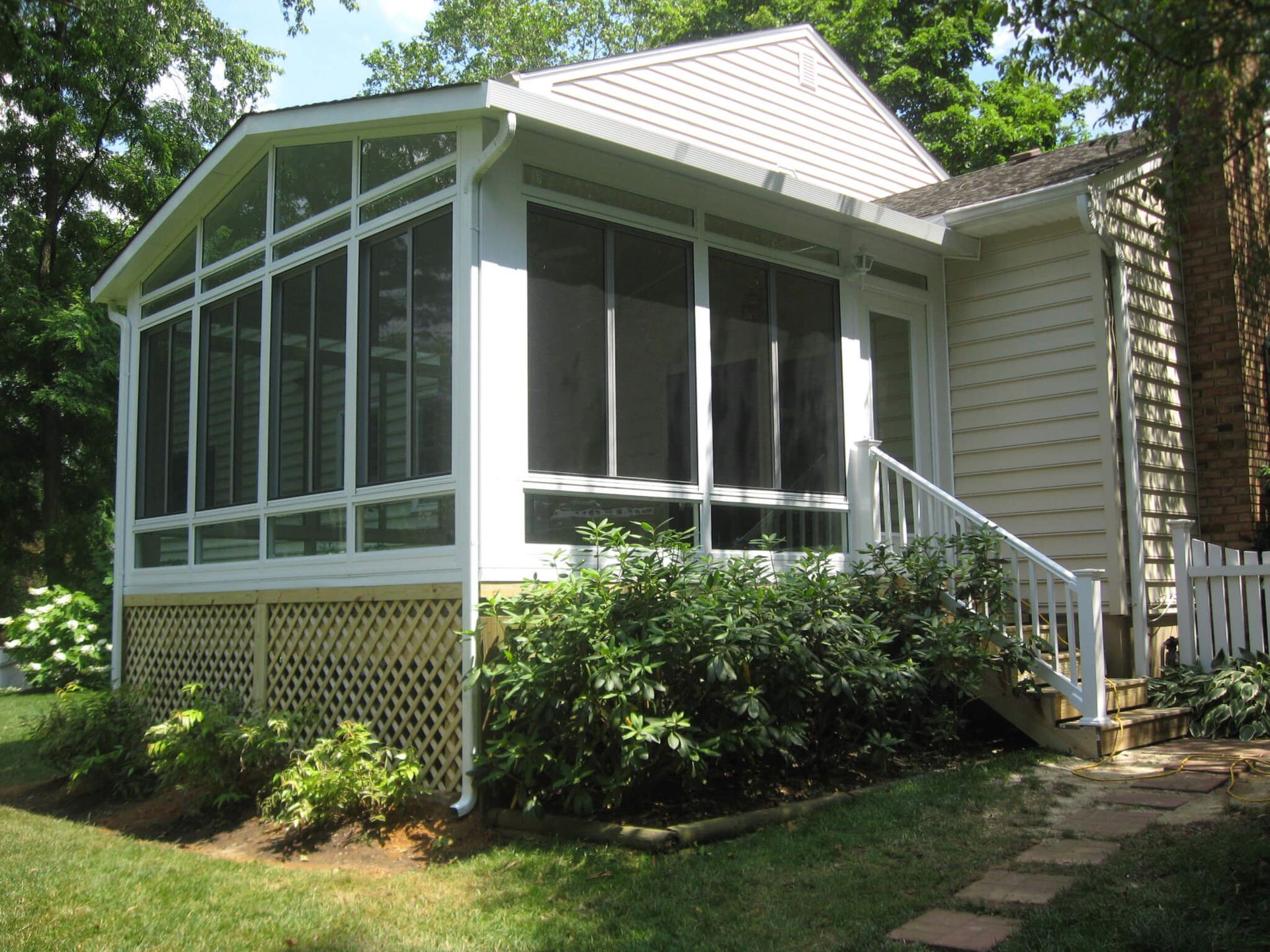 Screened-in porch addition with white trim, latticework, and small entry stairs. Green bushes and grass surround it.