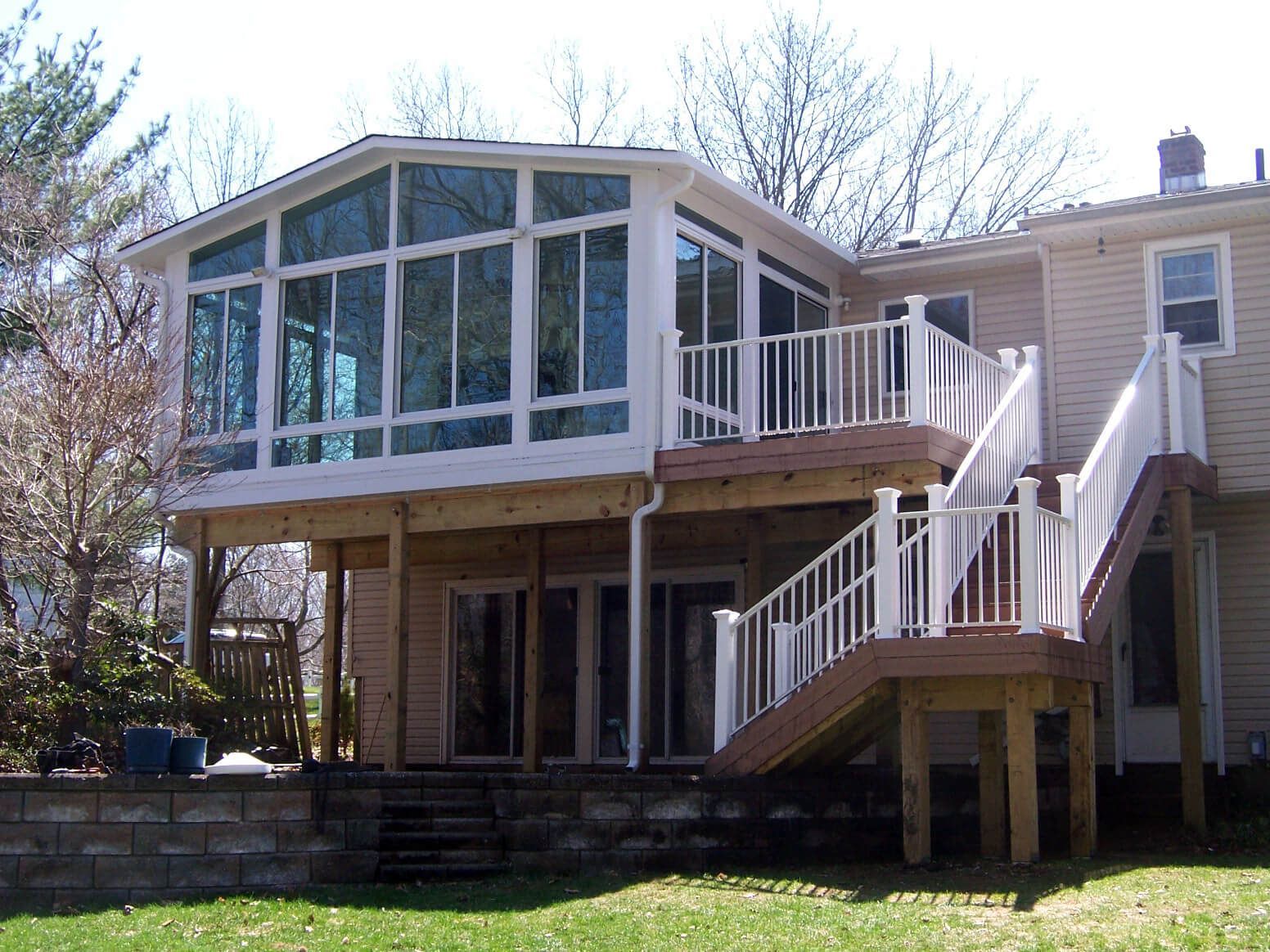 Two-story deck with sunroom, white railings, and stairs attached to a beige house, with a green lawn.