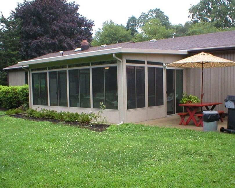 A backyard with a screen room, picnic table, umbrella, and grill. Green grass and trees are in the background.