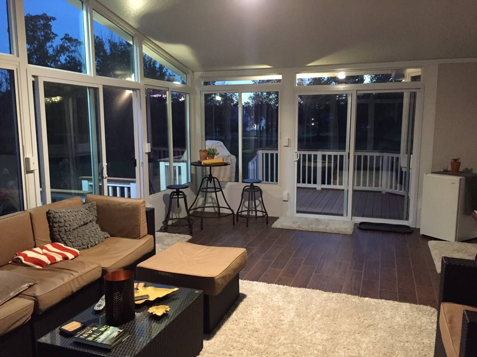 Sunroom with brown sofa, stools, and a table. Large windows overlook a dark outdoor scene.