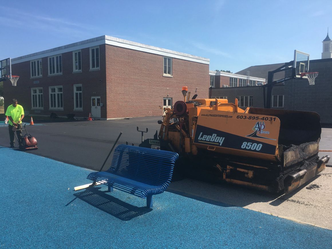 Asphalt paving underway near a school building; blue bench and worker in high-vis vest visible.