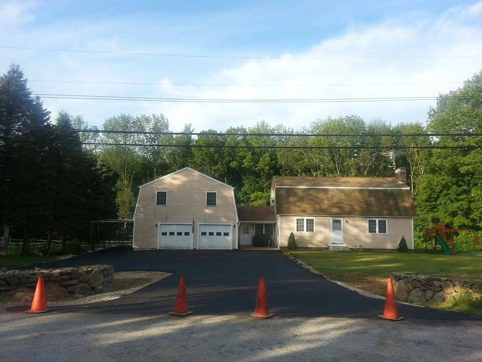 Driveway leading to house and garage, blocked by orange cones. Trees in background, blue sky.
