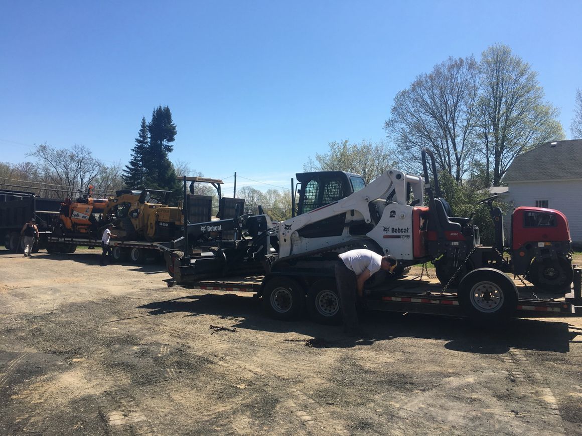 Construction equipment on trailers outdoors, under a blue sky; a person works on the machinery.