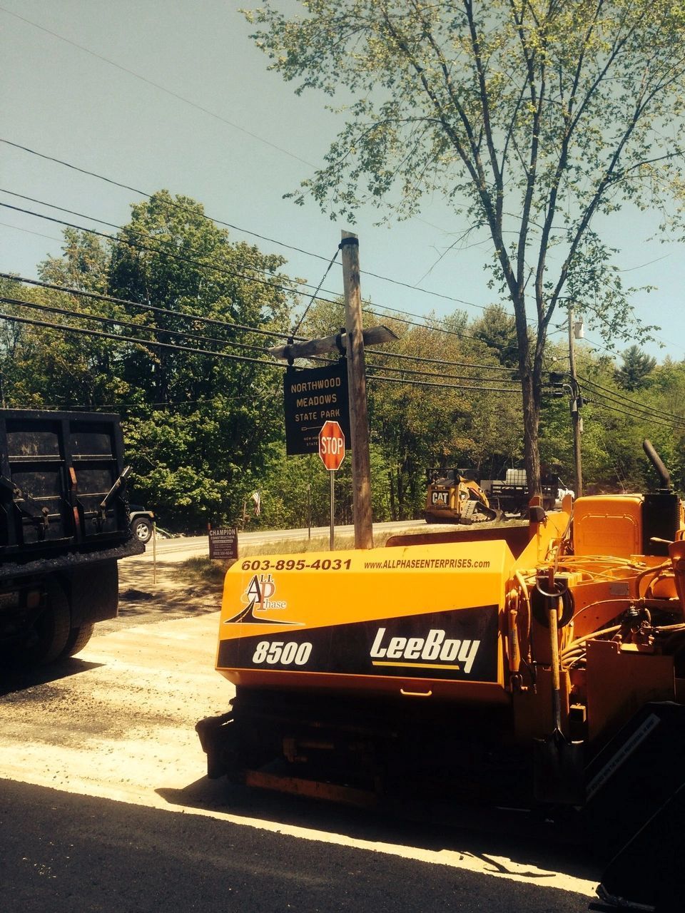 Orange LeeBoy asphalt paver on road with dump truck, trees, and utility pole.