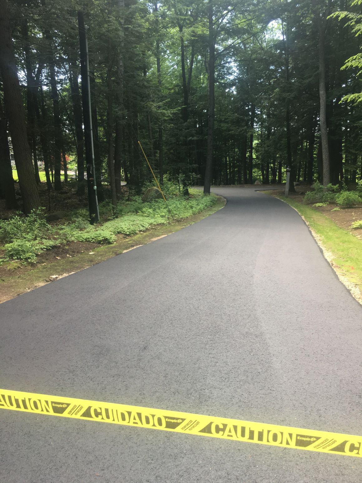 Newly paved asphalt road through a wooded area, blocked by yellow caution tape.