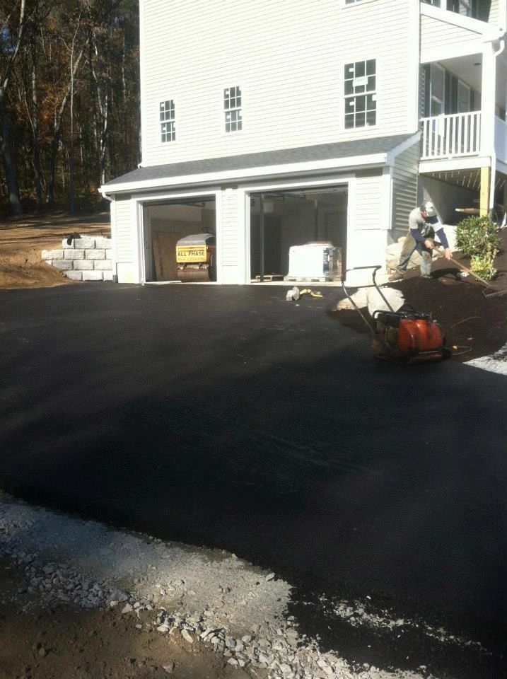 Asphalt driveway being rolled; garage and house in background. Person near garage, working.