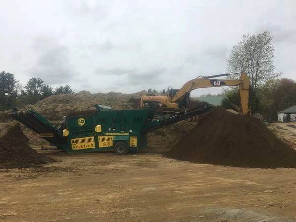 An excavator loading soil onto a green soil screener. Piles of soil are in a construction setting, under a cloudy sky.
