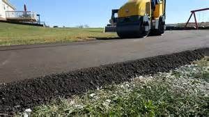 Yellow road roller compacting fresh asphalt on a pathway, grass in foreground, residential setting.