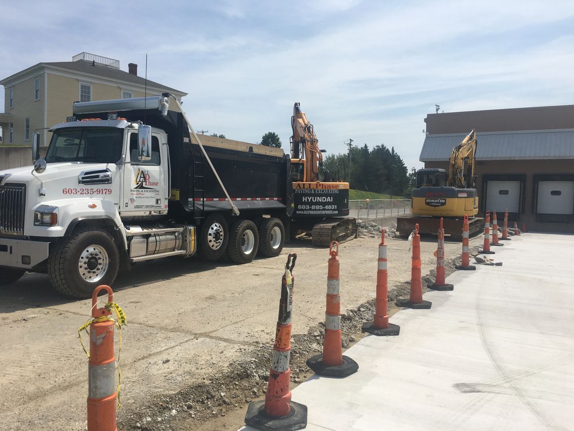 Construction site: Dump truck and excavator near building with loading docks, separated by traffic cones.