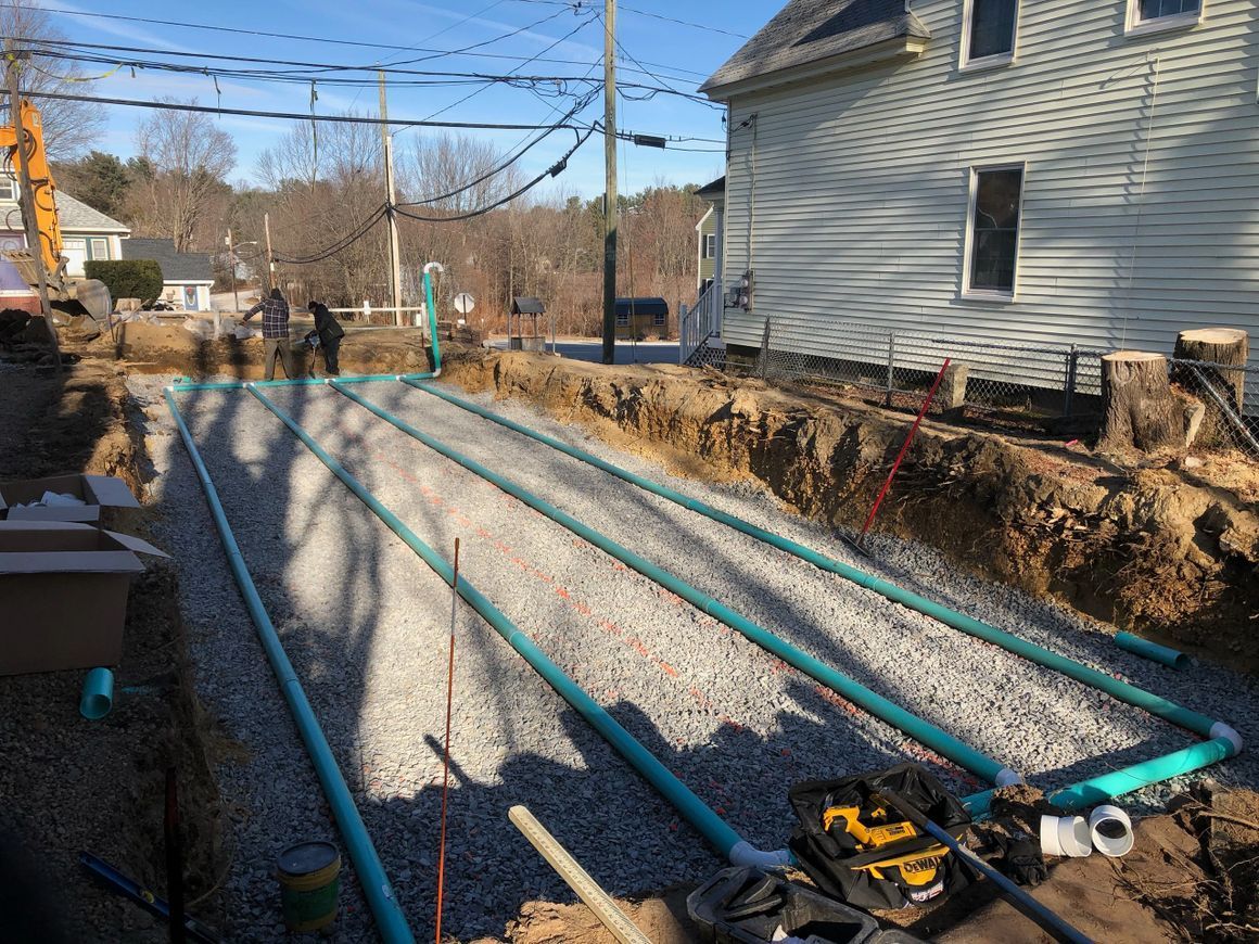 Trenches with gravel and blue pipes being installed for a septic system near a house.