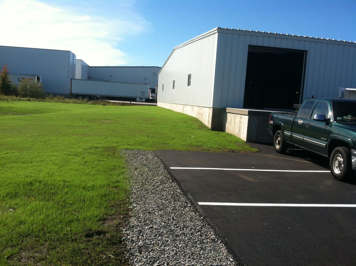 Green pickup truck parked near a large, white industrial building with a loading dock and grassy area.