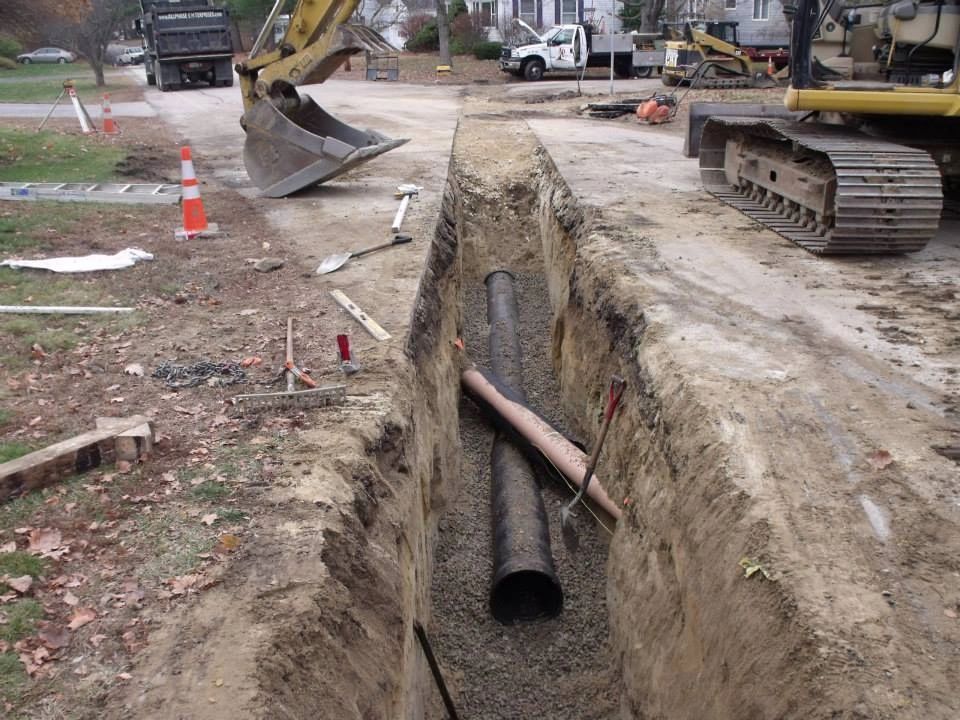 Construction site with an open trench containing pipes, alongside heavy machinery and a street.