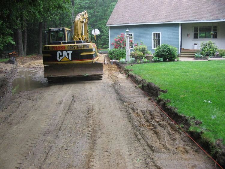 An excavator clearing a muddy driveway next to a house with a green lawn.