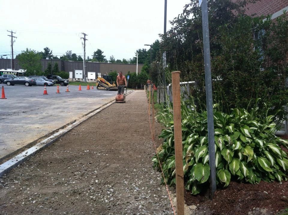 Person operating a vibrating plate compactor on a gravel pathway next to a building and parking lot.