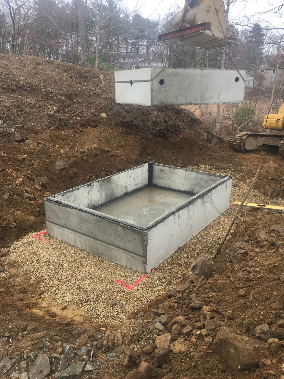 Concrete septic tank components being installed in a gravel-lined excavation pit. A backhoe is visible.