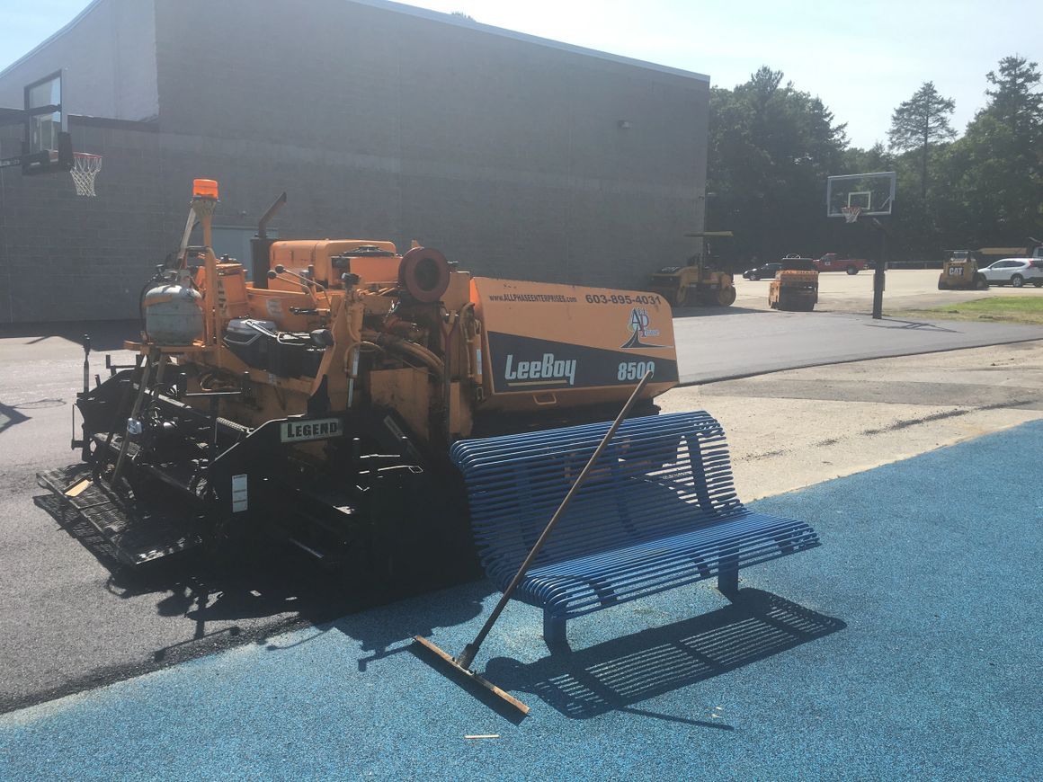 Asphalt paving machine on blacktop near blue bench and basketball court.