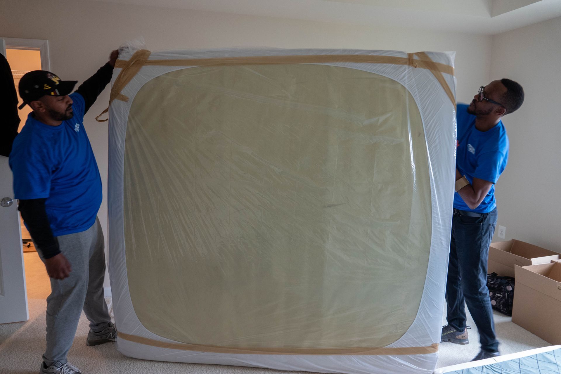 Two people in blue shirts lift a large, wrapped mattress in a room.