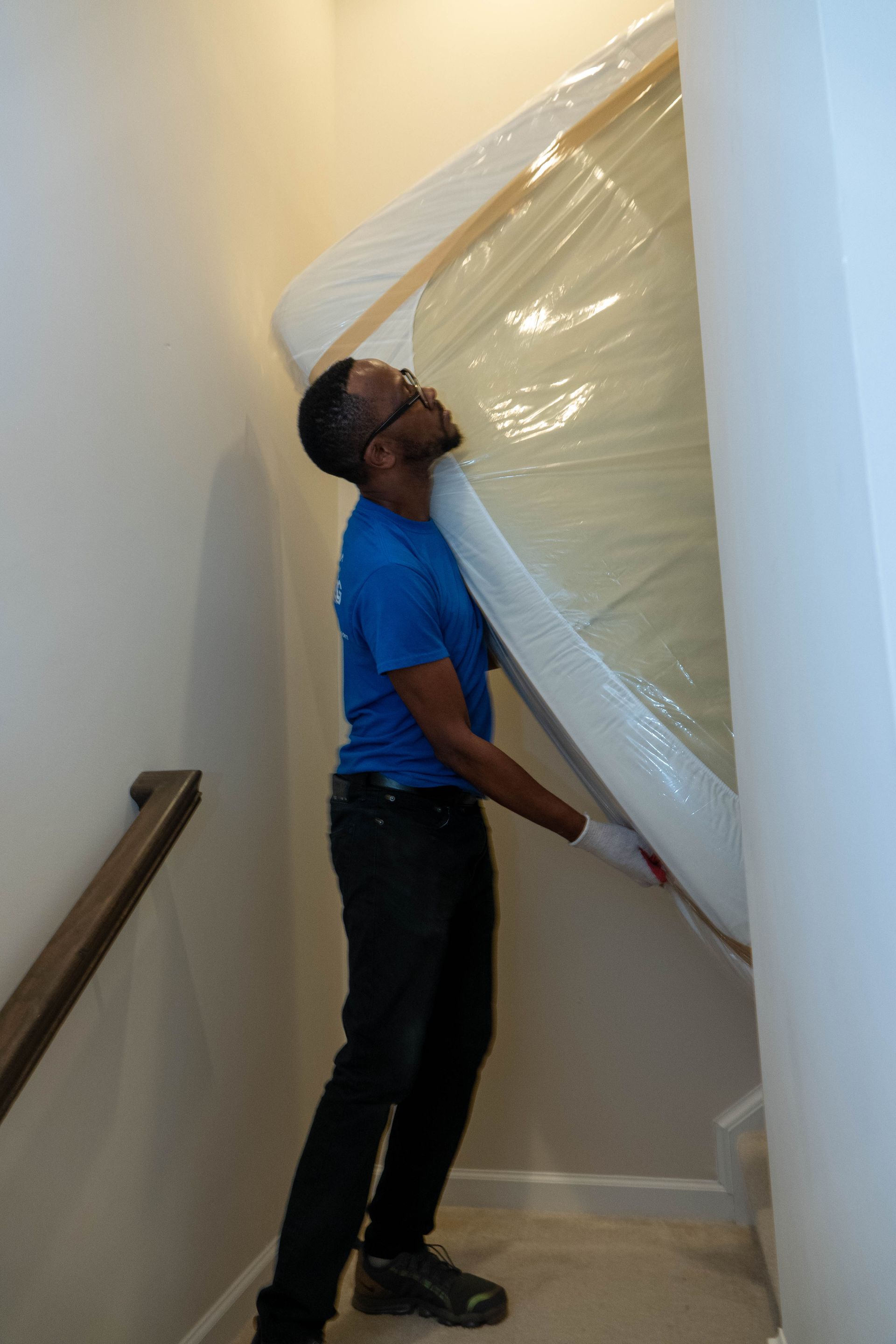 Man carrying a large, wrapped mattress up a narrow flight of stairs.
