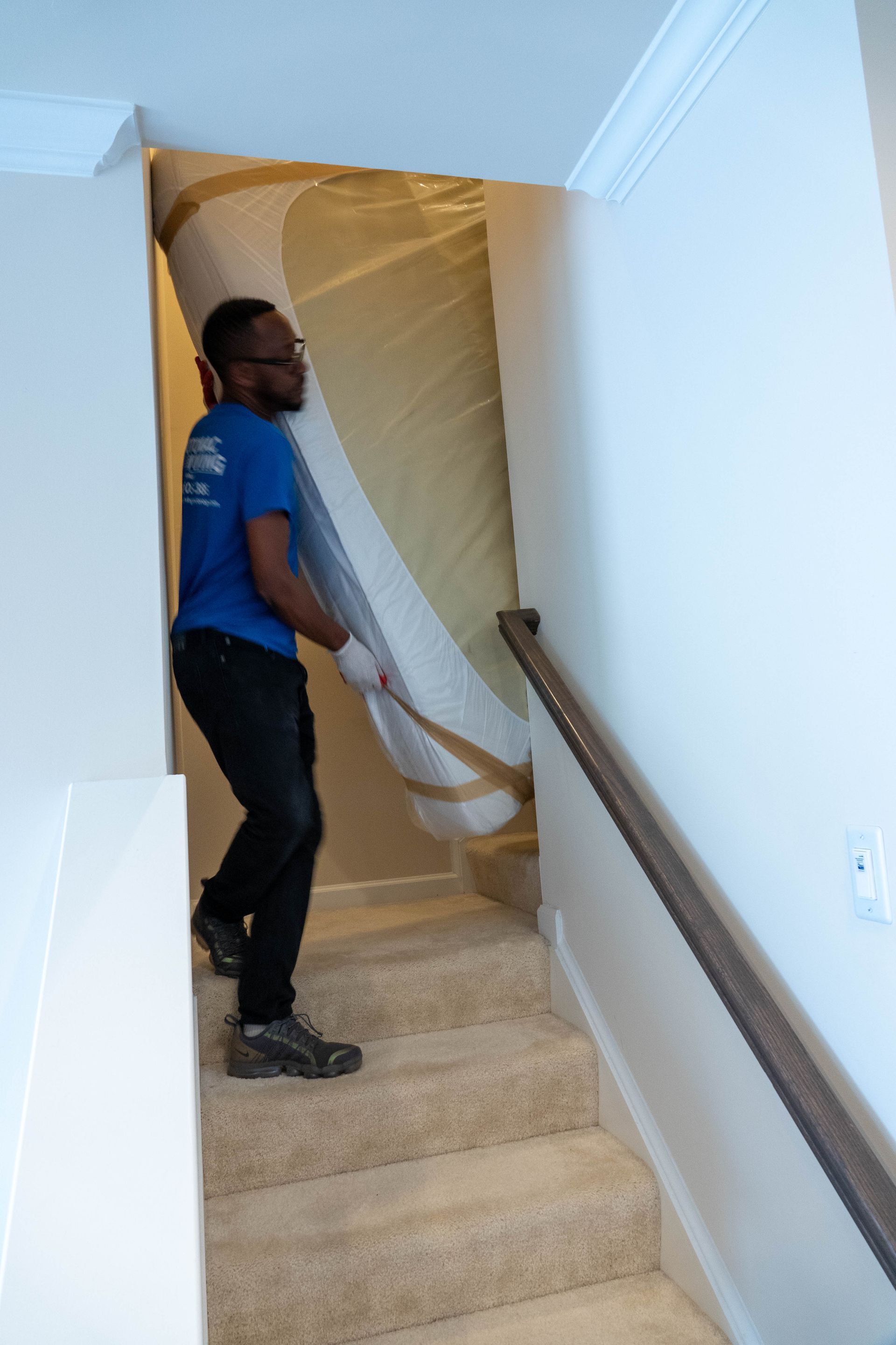 A person carrying a large, wrapped mattress up a carpeted staircase with a wooden handrail.