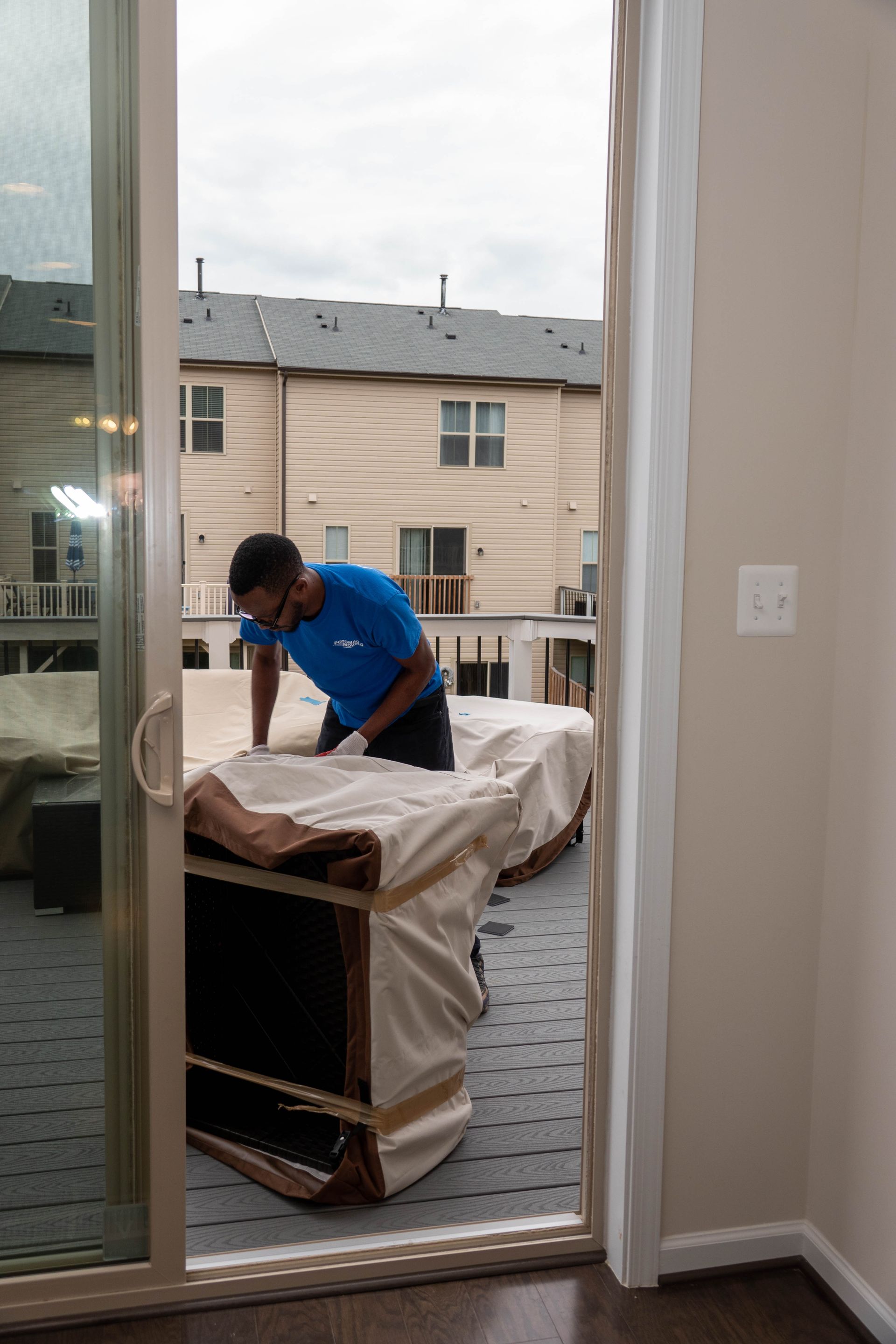 Person in blue shirt moving a wrapped sofa through a sliding glass door onto a patio, houses in background.