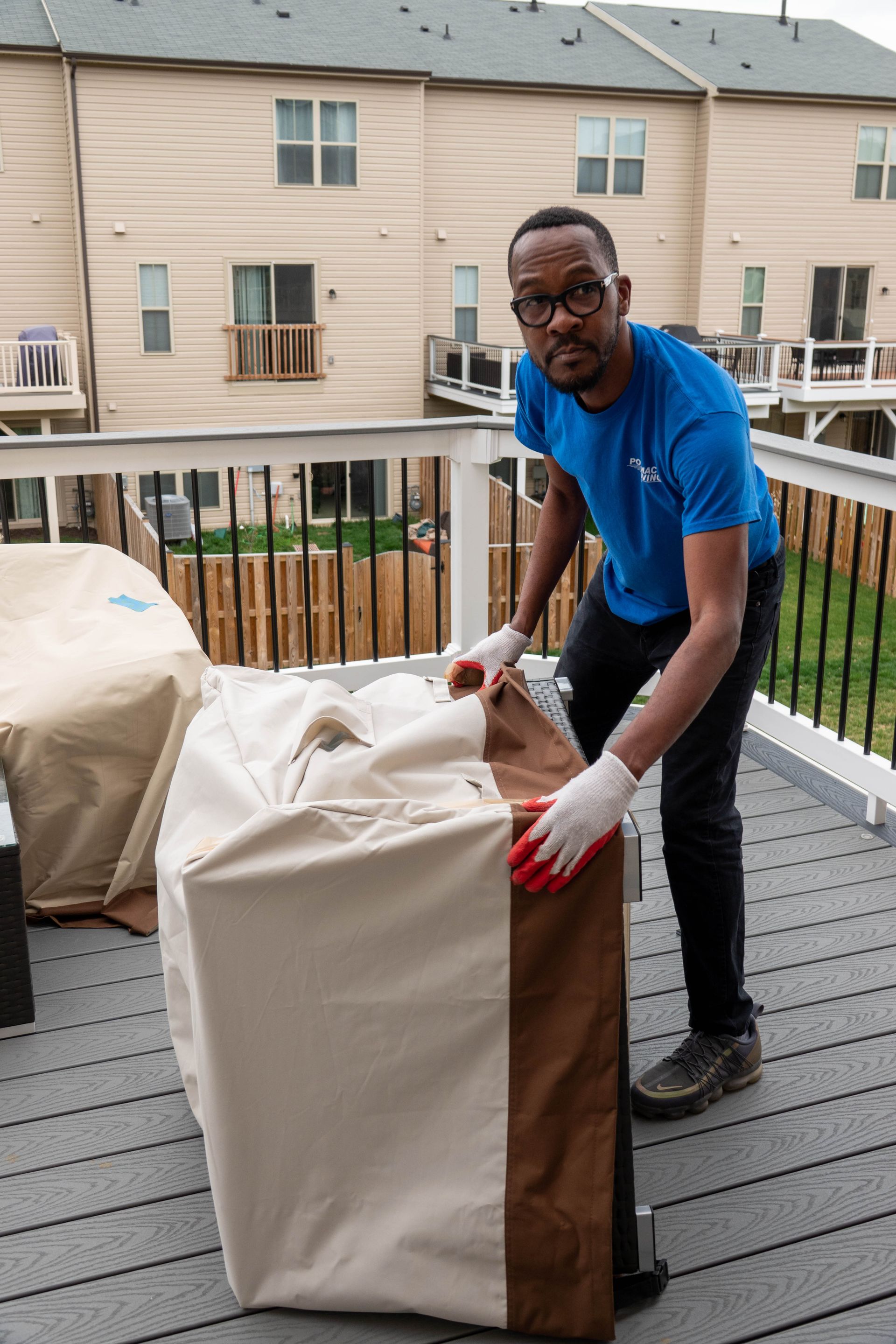 Person on a deck putting a cover on a cabinet. Wearing a blue shirt, black pants, and gloves. Beige cover.