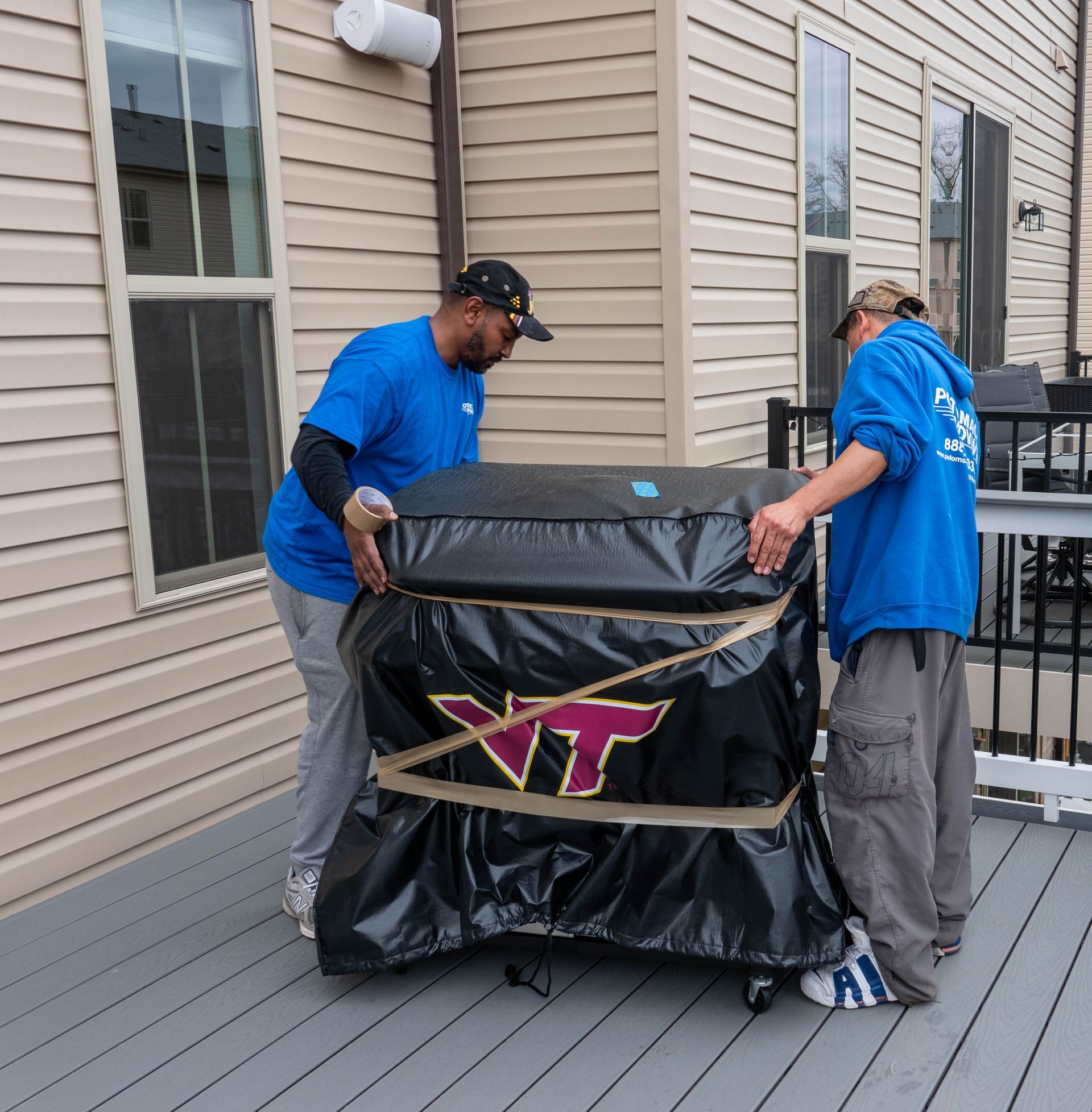 Two men in blue shirts lift a covered object with a VT logo on a deck.