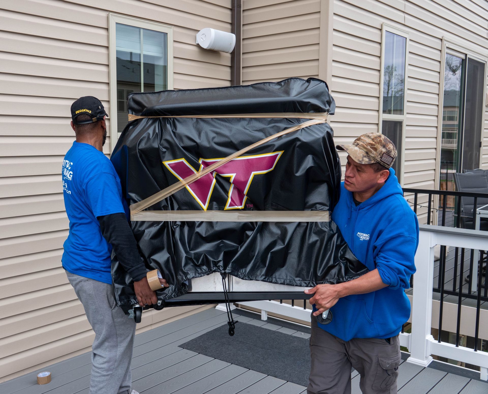 Two people carrying a large object wrapped in black tarp with a VT logo on a wooden deck.