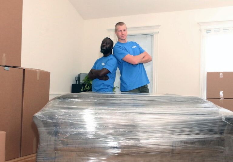 Two movers in blue shirts lean against each other, arms crossed, behind a wrapped sofa. Boxes in a room.