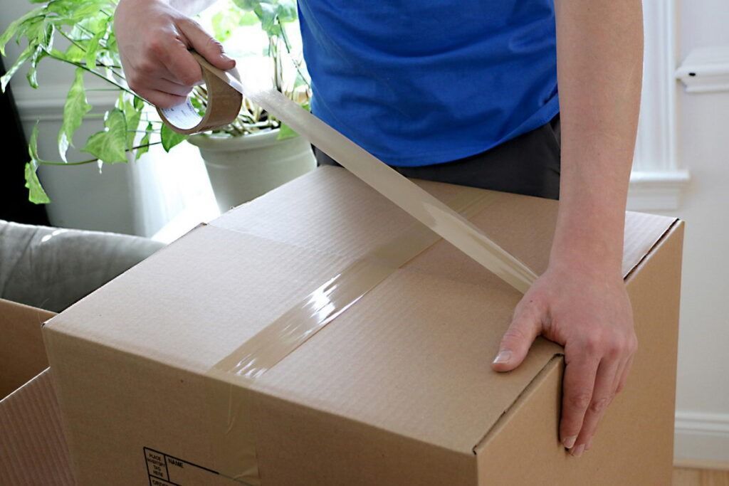 Person taping a cardboard box shut indoors, preparing for a move.