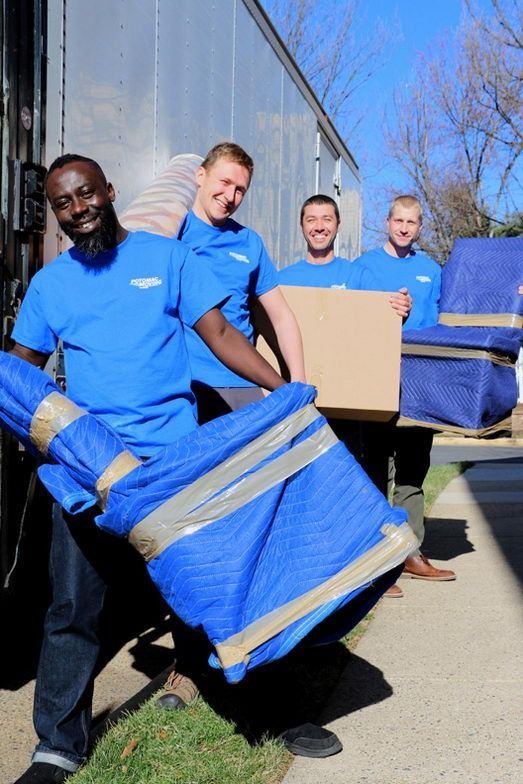 Four movers in blue shirts carrying wrapped furniture, a box, and smiling outside a truck.