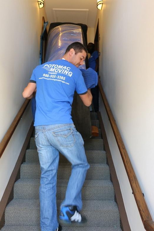 Movers carrying a large, wrapped item up a stairwell. Blue shirt, jeans, wooden stairs.