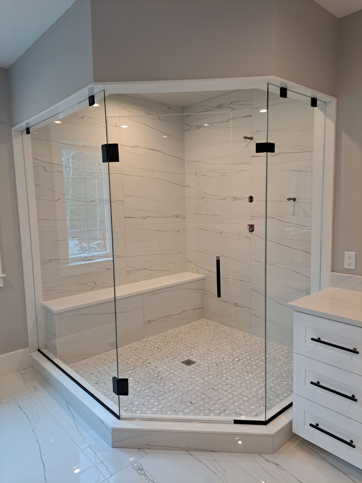 A bathroom with a walk in shower with a bench and a window.