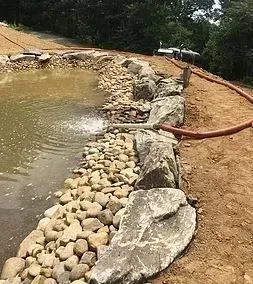 A pond with a rocky edge and a water fountain, with construction materials and a hose nearby.