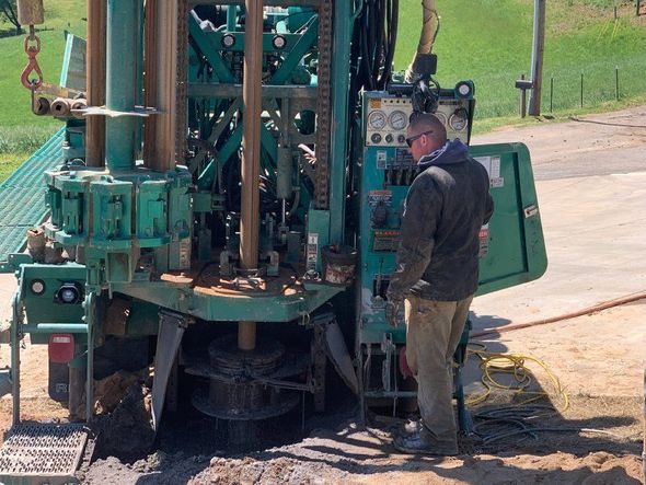 Person operating a large green drilling rig in an outdoor setting, drilling into the ground.