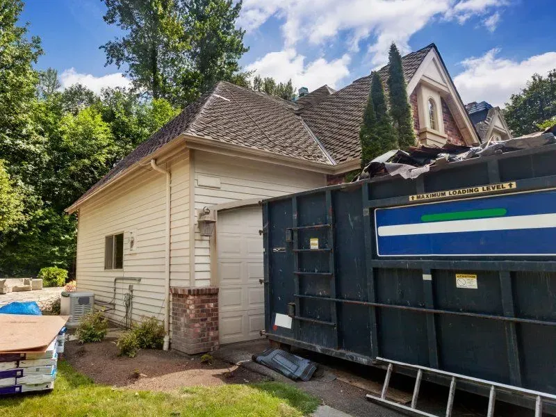 Garage beside a house with a dumpster in the driveway; roof repair underway.
