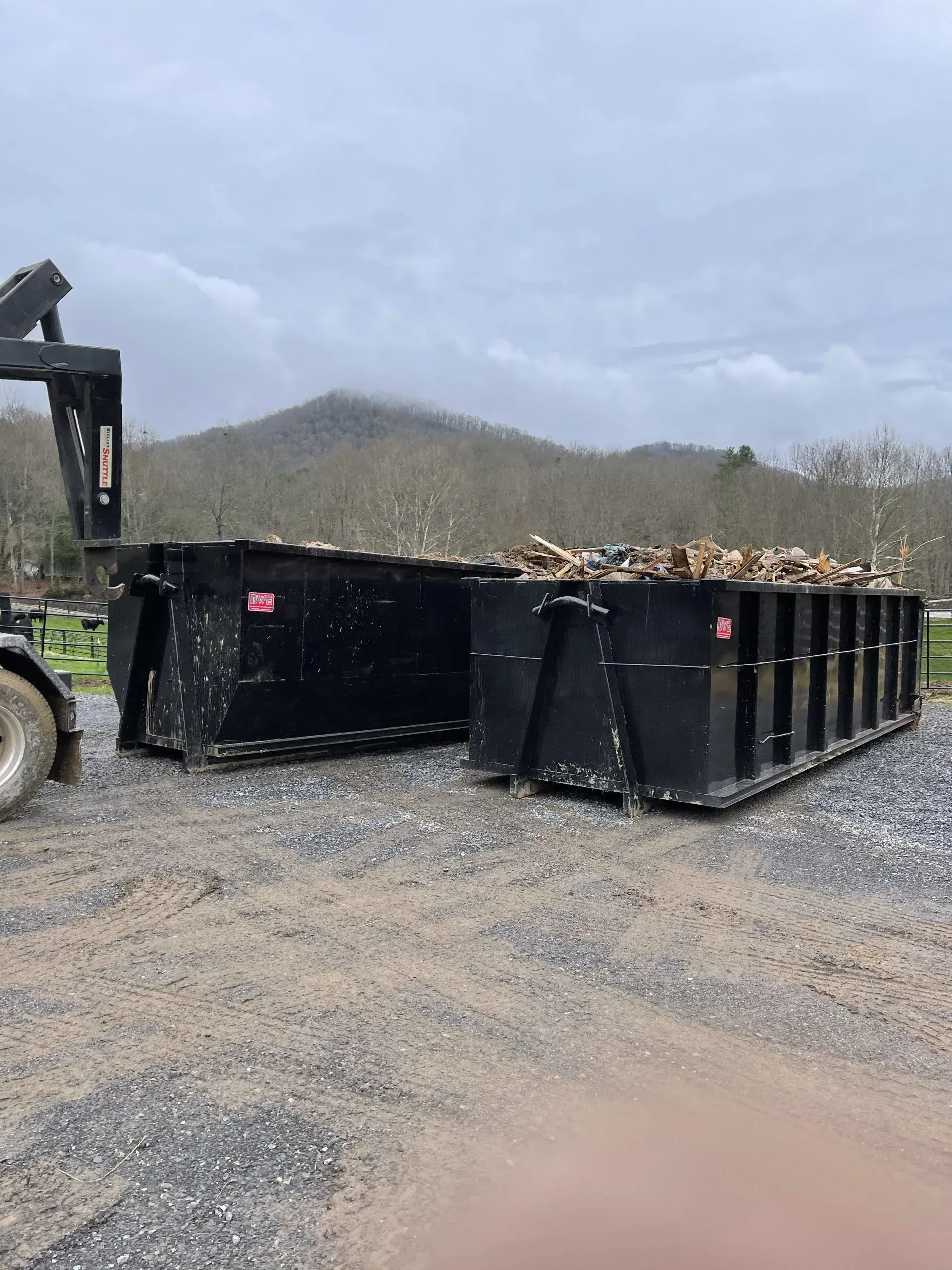Two black dumpsters filled with wood debris, parked on gravel with a foggy mountain background.