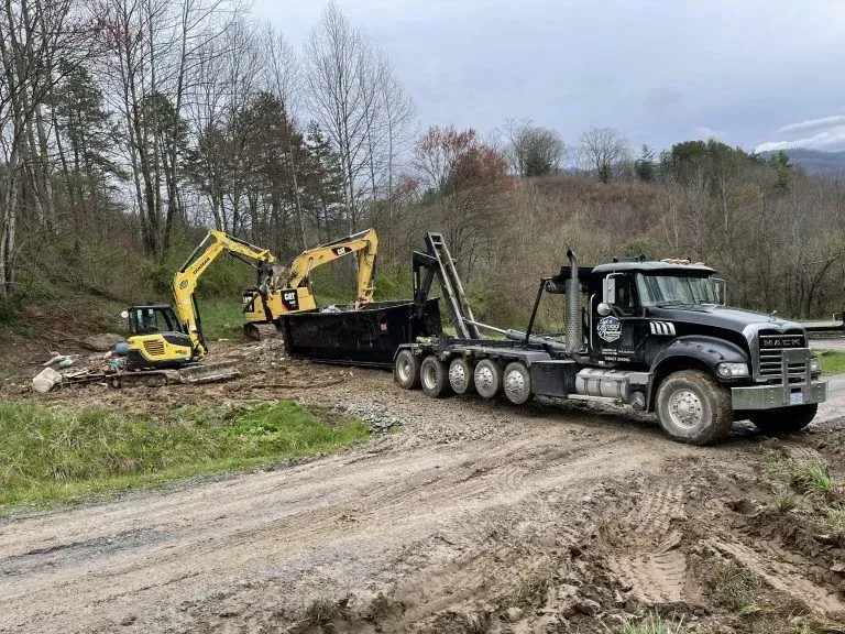 Black truck with container and yellow excavators on muddy road in wooded area.