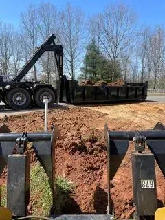 A construction scene: a dump truck with a full container is next to a hole filled with red soil.