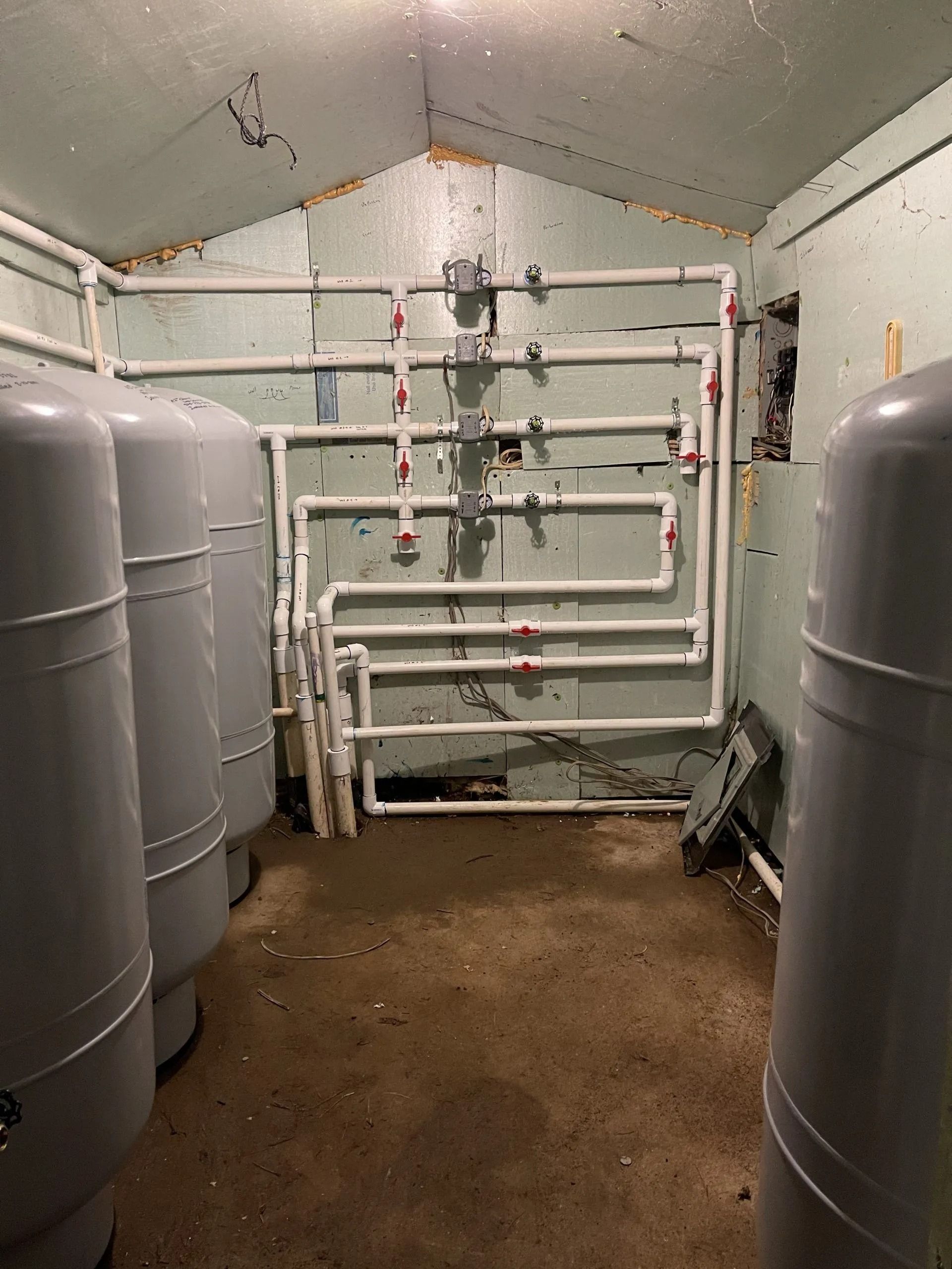 A small, indoor utility room with large gray tanks and white pipes along the walls. The floor is dirty.