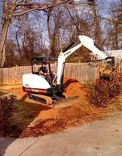 Mini excavator digging dirt near a fence. A person operates the machine outdoors.