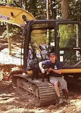 Man with dog sits on excavator in a wooded area. The man is wearing a hat, shirt, and shorts.