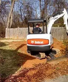A person operates a Bobcat excavator, digging in a yard near a wooden fence.