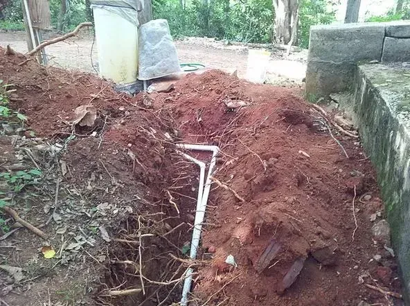 Trench dug in red soil, revealing white PVC pipe. Dirt pile beside stone structure and trees.