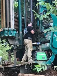 Man working on a drilling rig outdoors, wearing work clothes. Turquoise machinery and green foliage are visible.