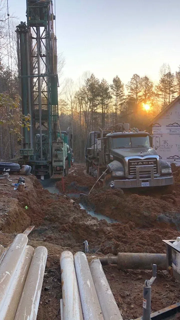 Drilling rig and truck on a muddy construction site at sunset, with plastic pipes in foreground.